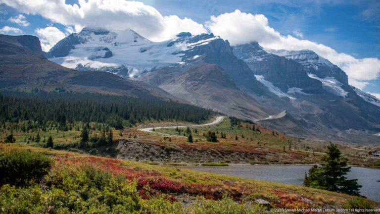 A glacier- and snow-covered mountain rises above an alpine pond, conifers, and red-gold low vegetation a road curves through the middle distance.