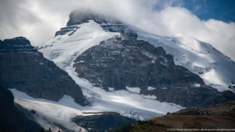 A close view shows snow and glacier ice draped across a steep, horizontally layered mountain beneath low cloud.