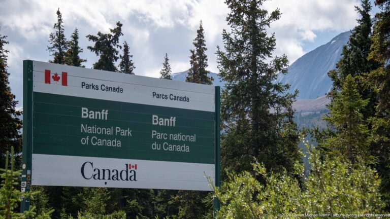 A green-and-white bilingual Parks Canada sign reads Banff National Park / Parc national du Canada among conifers.