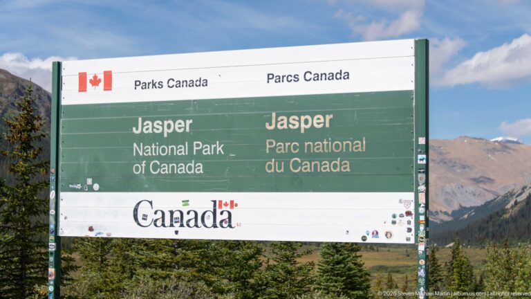A green-and-white bilingual Parks Canada sign reads Jasper National Park / Parc national du Canada with mountains behind.
