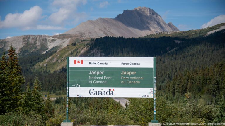The Jasper National Park sign stands low in the frame before conifer forest and a sharply profiled rocky mountain.