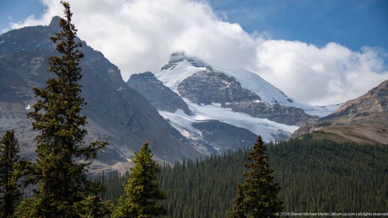 A glacier and snowfield descend from a cloud-wrapped summit between dark rock walls, with conifers in the foreground.