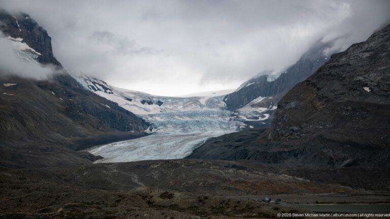 A broad glacier descends between dark mountain walls beneath low cloud, with gray moraine and forefield terrain in front.