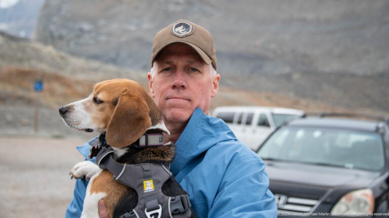 An adult in a blue jacket holds a beagle in a harness parked vehicles and a barren mountain slope are softly focused behind.