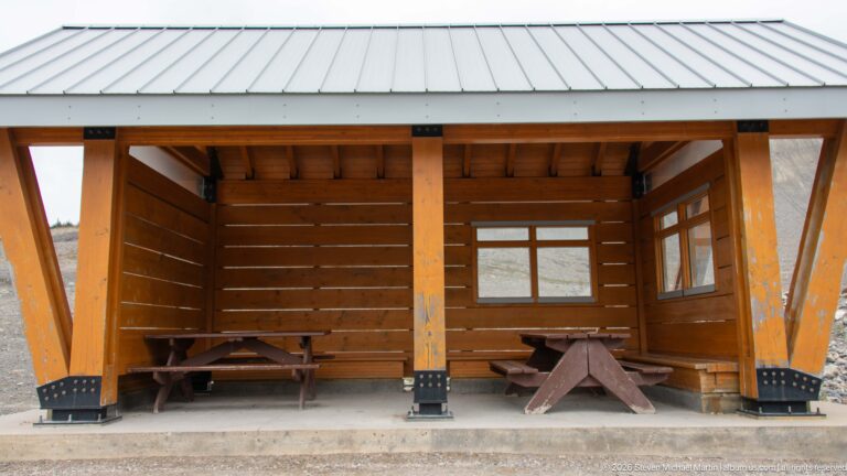 An open timber shelter with metal roof contains picnic tables and benches on a concrete pad.