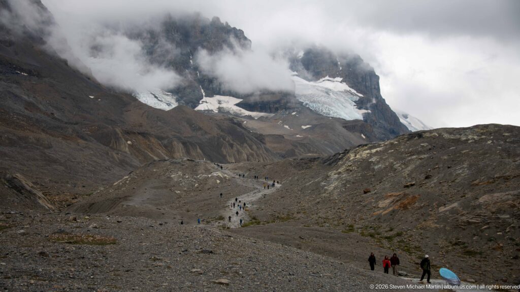 Numerous small figures follow a pale trail across barren moraine toward glacier ice beneath low cloud and steep rock walls.