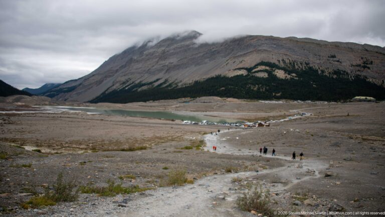 A broad barren valley floor with a pale trail and scattered visitors extends toward water and mountain slopes under overcast skies.