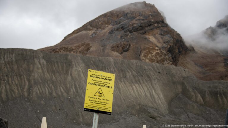 A yellow natural-hazards warning sign stands before steep gray moraine and a cloud-wrapped rocky peak.