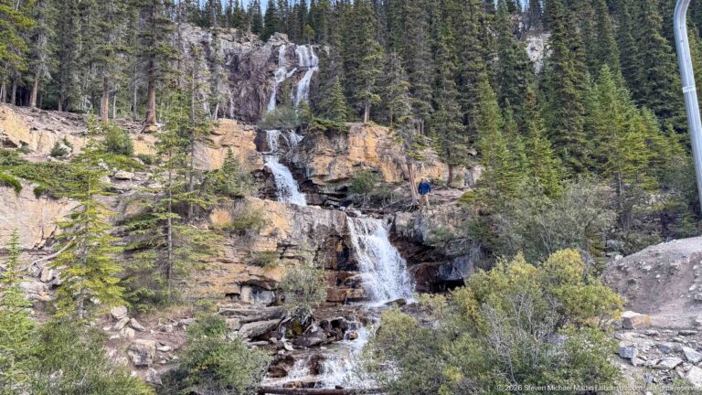 A narrow waterfall drops over layered rock ledges beside conifers, shrubs, and a visible visitor for scale.