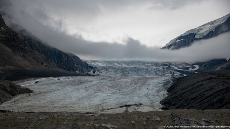 Glacier ice fills the valley between dark moraine slopes and steep mountains, with low cloud obscuring the upper accumulation area.