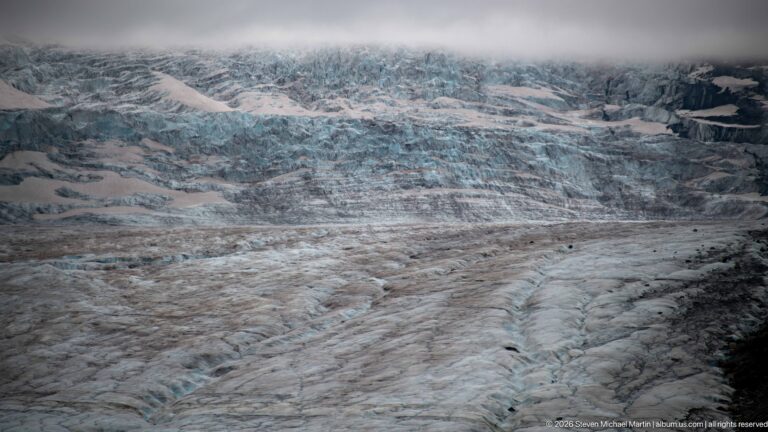 A tight view shows fractured blue-white glacier ice crossed by gray debris bands, ridges, and shallow crevasses.