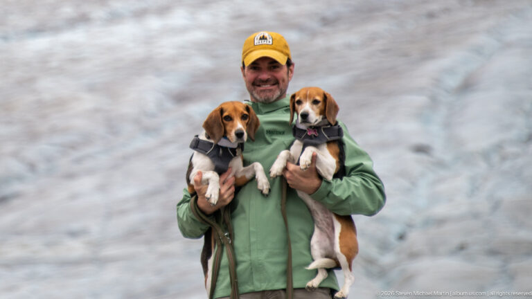 A smiling adult holds two beagles in harnesses the glacier surface forms a softly focused blue-gray background.