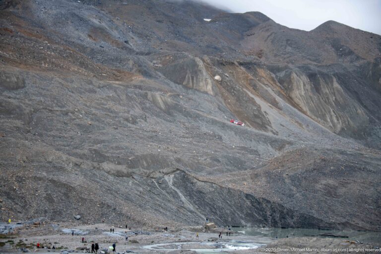 A vast barren gray-brown slope is cut by erosion channels and pale sediment bands tiny visitors and a red vehicle provide scale near the base.