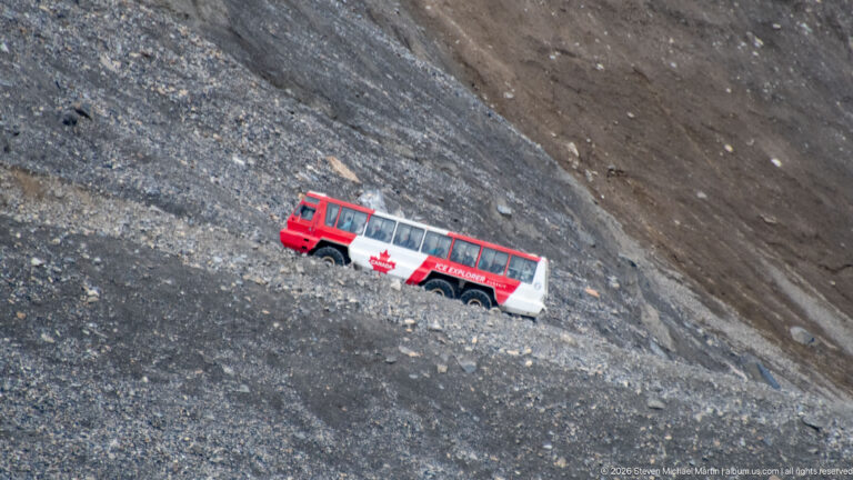 Red-and-white Ice Explorer tour vehicle crossing a steep gray-brown rocky moraine slope.