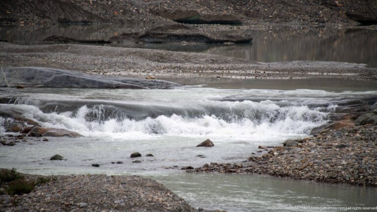 Pale glacial meltwater cascades over low rock ledges through a gravelly barren forefield.