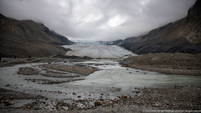 Athabasca Glacier beyond braided pale meltwater channels, gravel bars, and dark moraine slopes under low clouds.