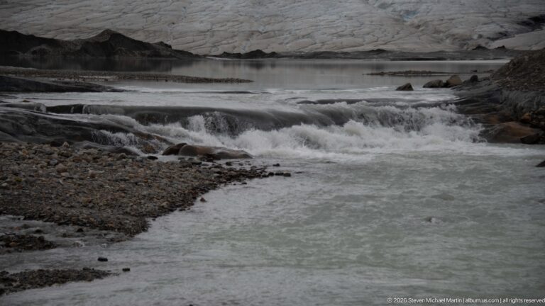 Milky glacial meltwater rushing over dark bedrock ledges with the edge of Athabasca Glacier beyond.