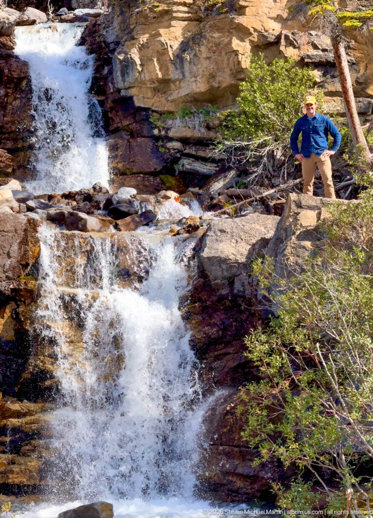 A visitor stands on a rock ledge beside foaming water descending through a rocky forest channel.