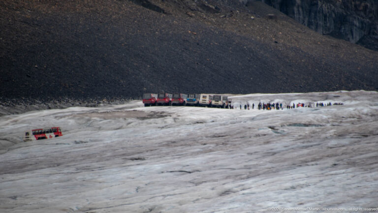 Red-and-white Ice Explorer vehicles and groups of visitors on Athabasca Glacier below a dark moraine slope.