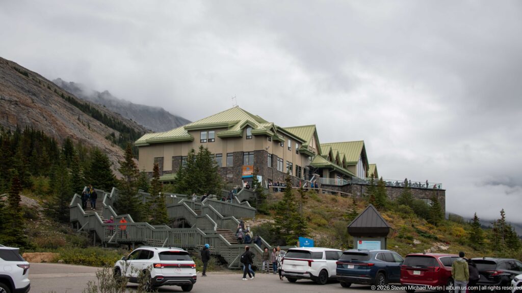 Columbia Icefield Glacier Discovery Centre with green roofs, stone walls, exterior stairs, parked cars, and visitors.