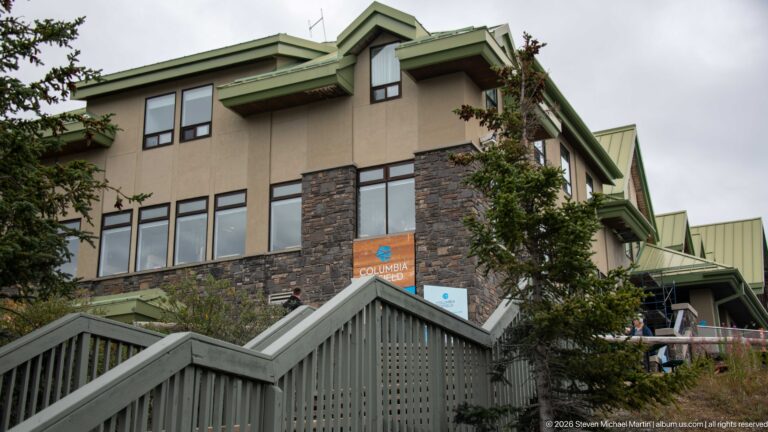 Glacier Discovery Centre exterior with stone walls, tan upper stories, green roofs, stairs, windows, and conifer trees.