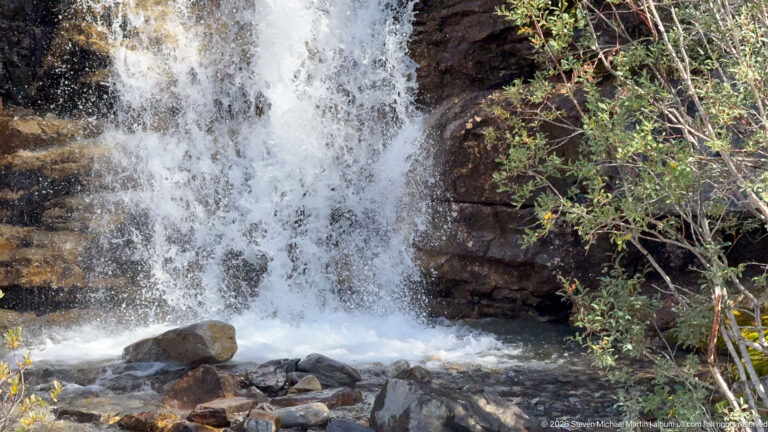 Foaming water drops over dark rock into a shallow boulder-filled pool, with leafy shrubs at right.