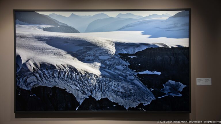 Large aerial glacier photograph showing a broad snowfield narrowing into heavily crevassed ice over dark mountain terrain.