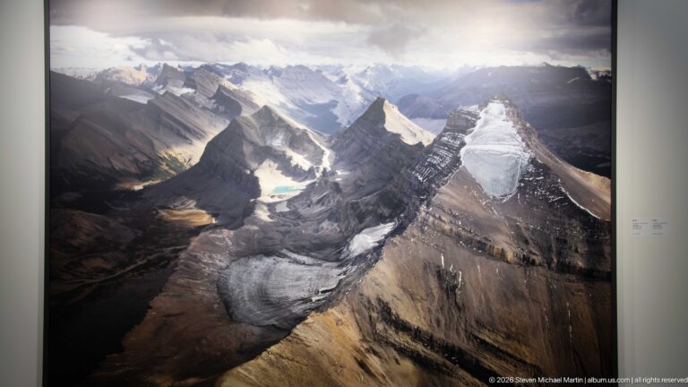 Large aerial photograph of steep mountain ridges, glaciers, turquoise lakes, and deep valleys beneath broken clouds.