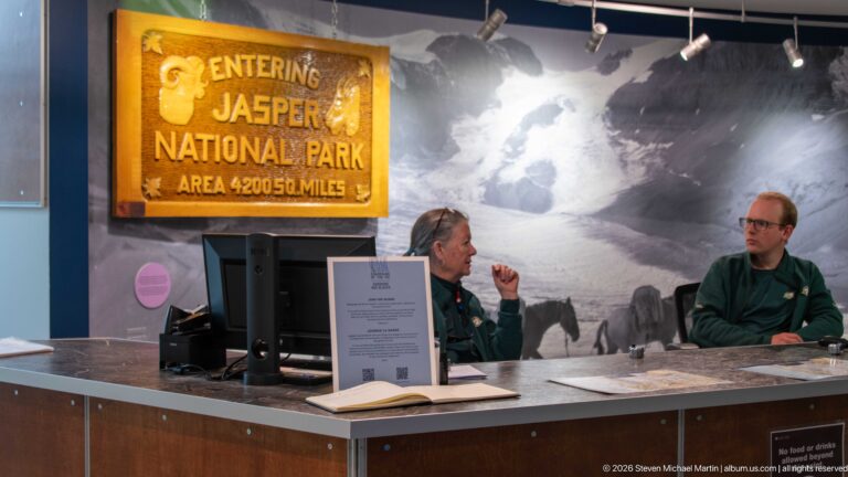 Two staff members at a visitor information counter beneath an Entering Jasper National Park sign and glacier mural.
