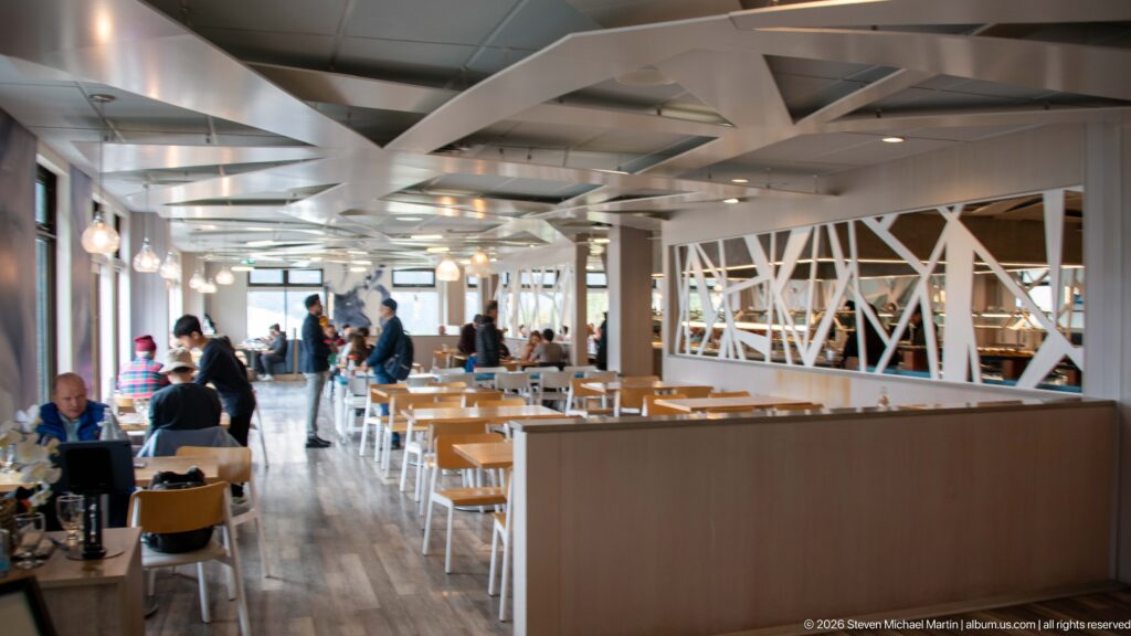Bright visitor-centre dining room with pale wood tables, white trusses, pendant lights, and seated visitors.