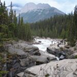 Turbulent Mistaya River crosses pale bedrock between conifer forests, with visitors along the rim and a mountain beyond.