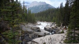 Turbulent Mistaya River crosses pale bedrock between conifer forests, with visitors along the rim and a mountain beyond.