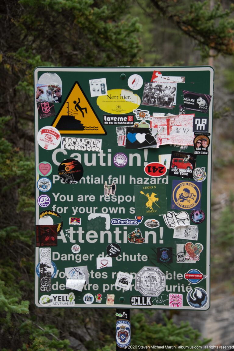 Sticker-covered warning sign beside the Mistaya Canyon route, with a yellow falling-person hazard symbol still visible.