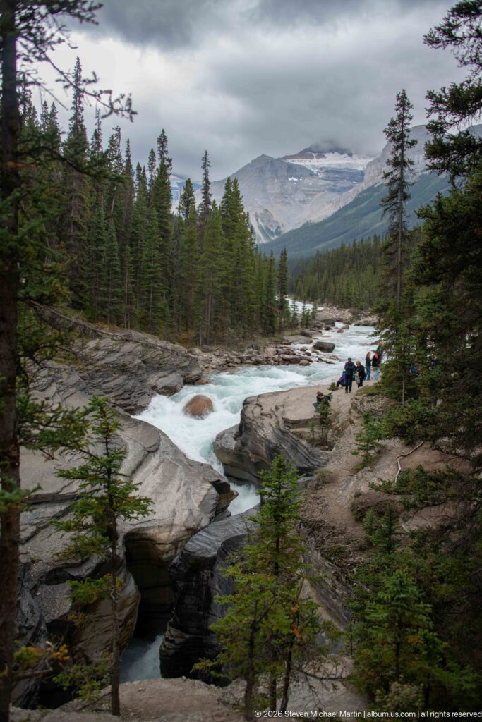 Whitewater on the Mistaya River crosses rock ledges above a dark canyon opening, backed by conifer forest and mountains.