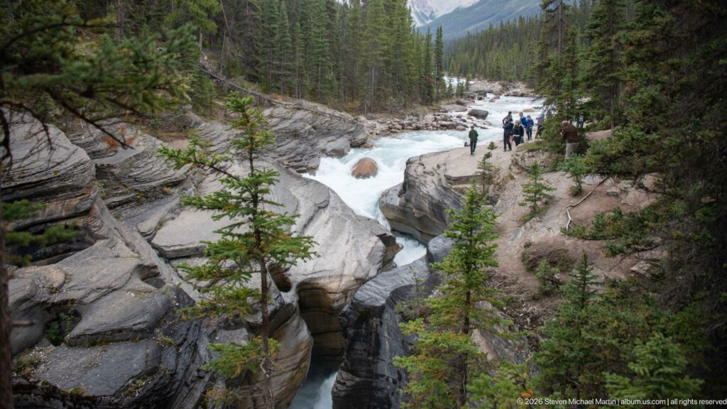 Narrow Mistaya River channel cuts between smooth rock walls, with rapids upstream, conifers, and visitors along the rim.