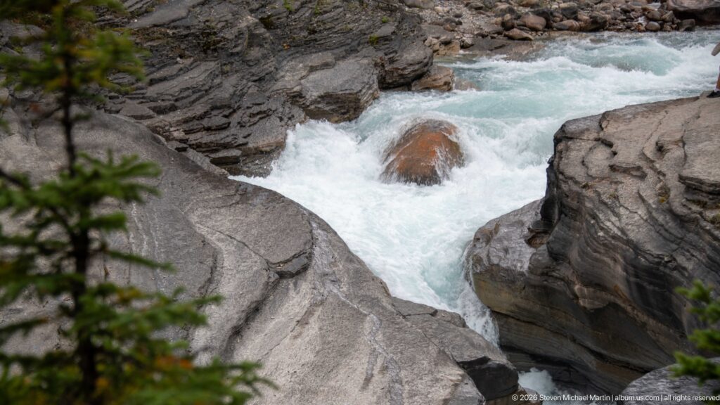 White Mistaya River rapids surge around a reddish boulder between smooth gray rock walls in the canyon.