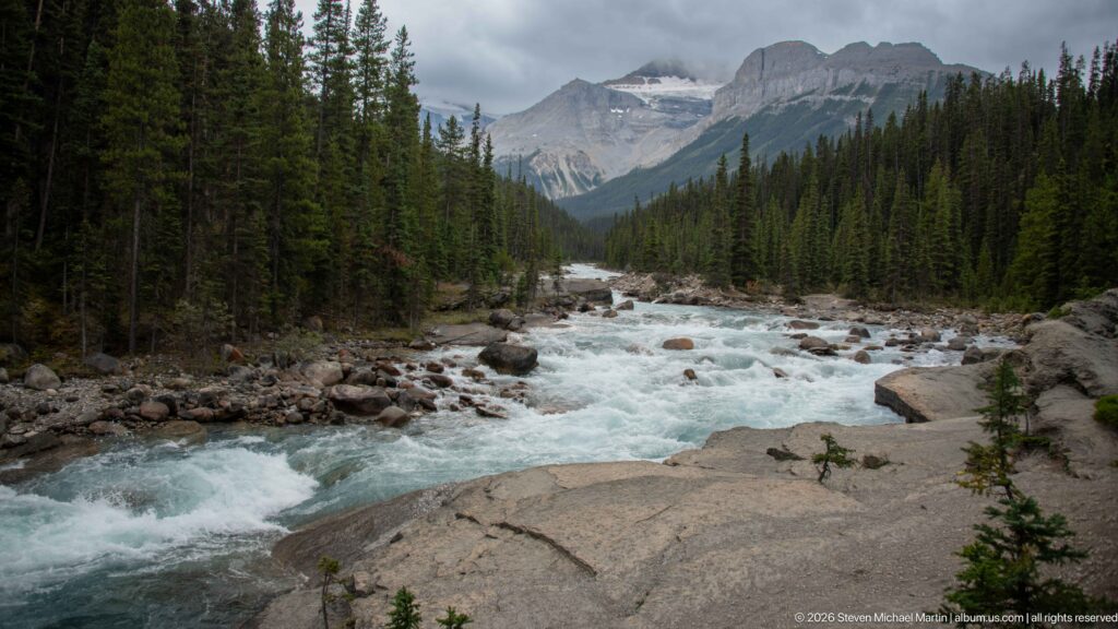 Fast blue-green river flows through a boulder-strewn channel between conifer forests, with steep mountains under low clouds.