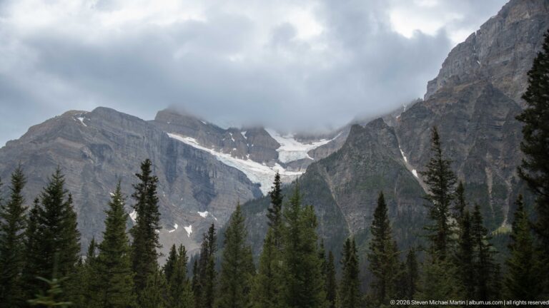 Glacier and snow fill a high basin between steep layered mountains above a foreground of dark conifers and low cloud.