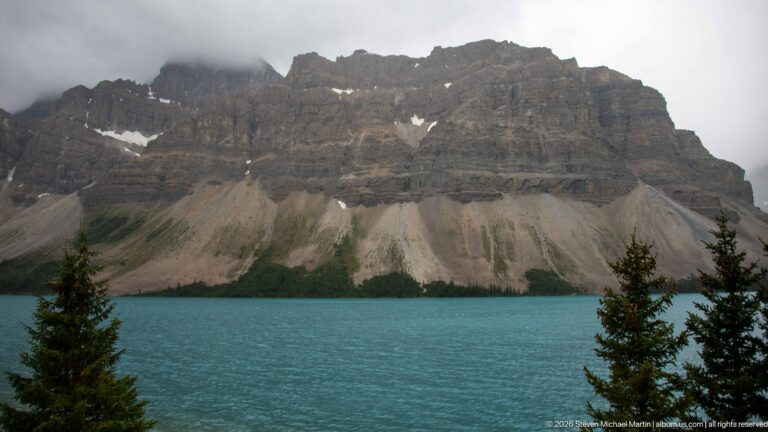 Turquoise mountain lake below layered cliffs and broad pale scree slopes, with conifers along the shoreline and low clouds above.