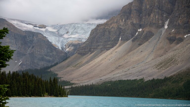 Glacier descends from a cloudy mountain basin above a turquoise lake, with dark conifer forest and broad scree slopes.