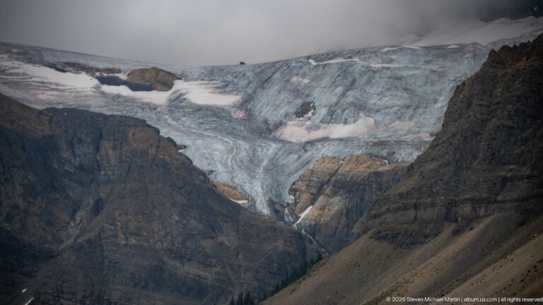Blue-gray crevassed glacier fills a high basin between dark rock walls, with snow patches, debris, and low cloud.