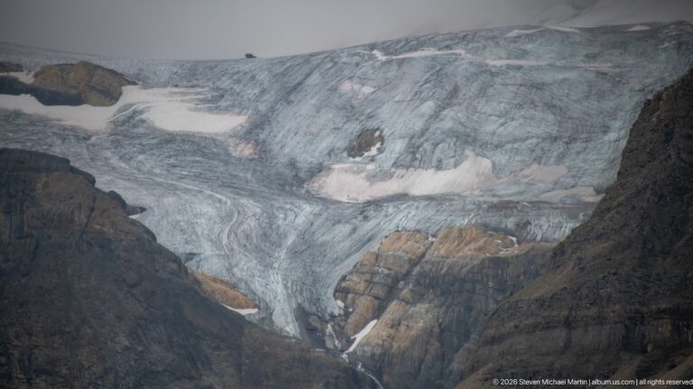 Close view of crevassed blue-gray glacier ice around exposed brown rock bands, with snow patches, debris, and a narrow meltwater streak.