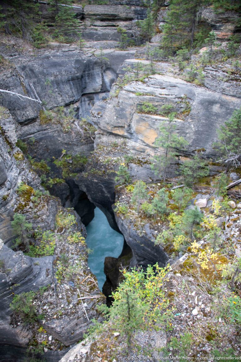 Blue-green river water appears through an eroded opening in broad gray bedrock surrounded by low vegetation and conifers.