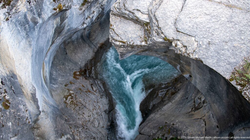 Turquoise Mistaya River water plunges through a narrow chute between smooth, pale rock walls in Mistaya Canyon.