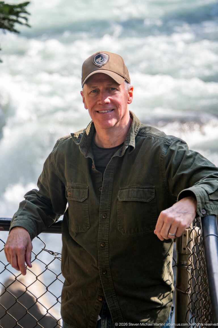Visitor leaning on a chain-link overlook railing with bright foaming river water behind him.