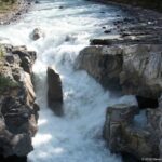 Powerful white water drops beside a tall central rock into a steep-walled gorge.