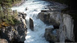 Powerful white water drops beside a tall central rock into a steep-walled gorge.