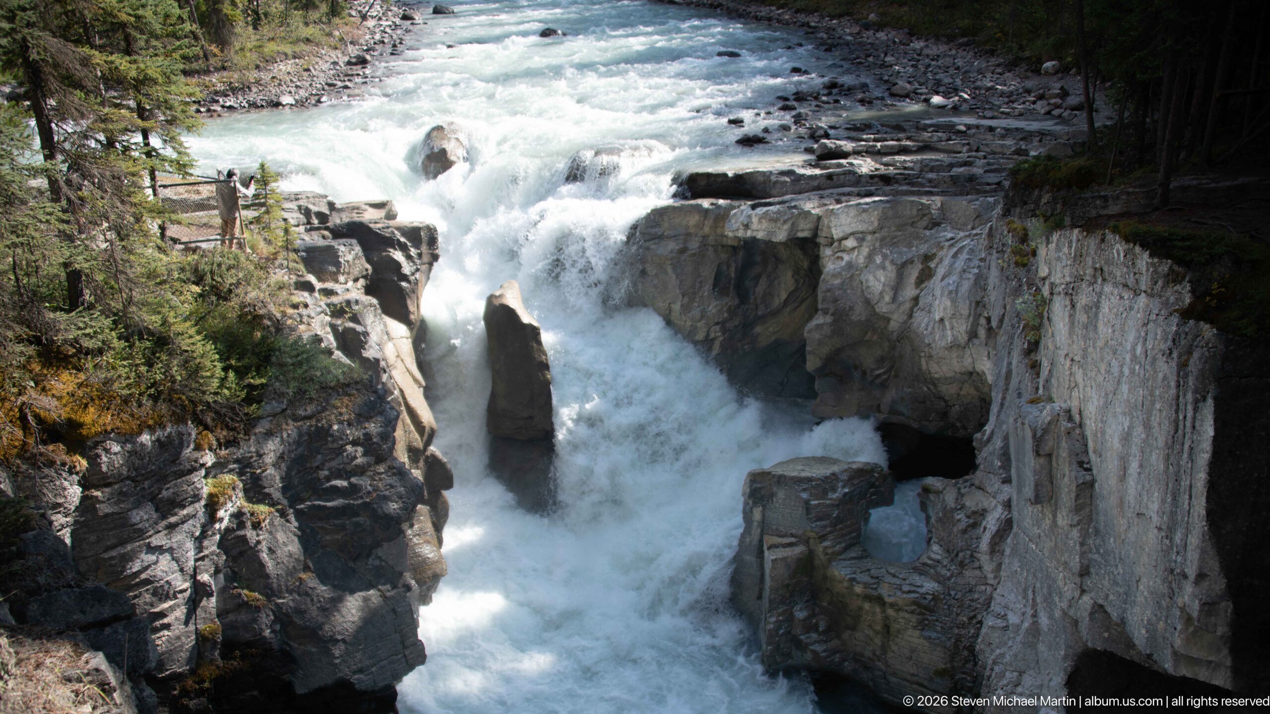 Powerful white water drops beside a tall central rock into a steep-walled gorge.
