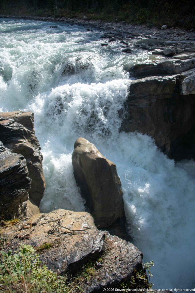 Close view of turbulent river water flowing around a large angular boulder and layered rock ledges.