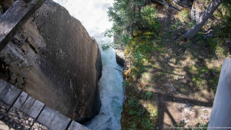 Steep downward view between rock walls toward white water, with overlook fencing and forest vegetation at the edges.